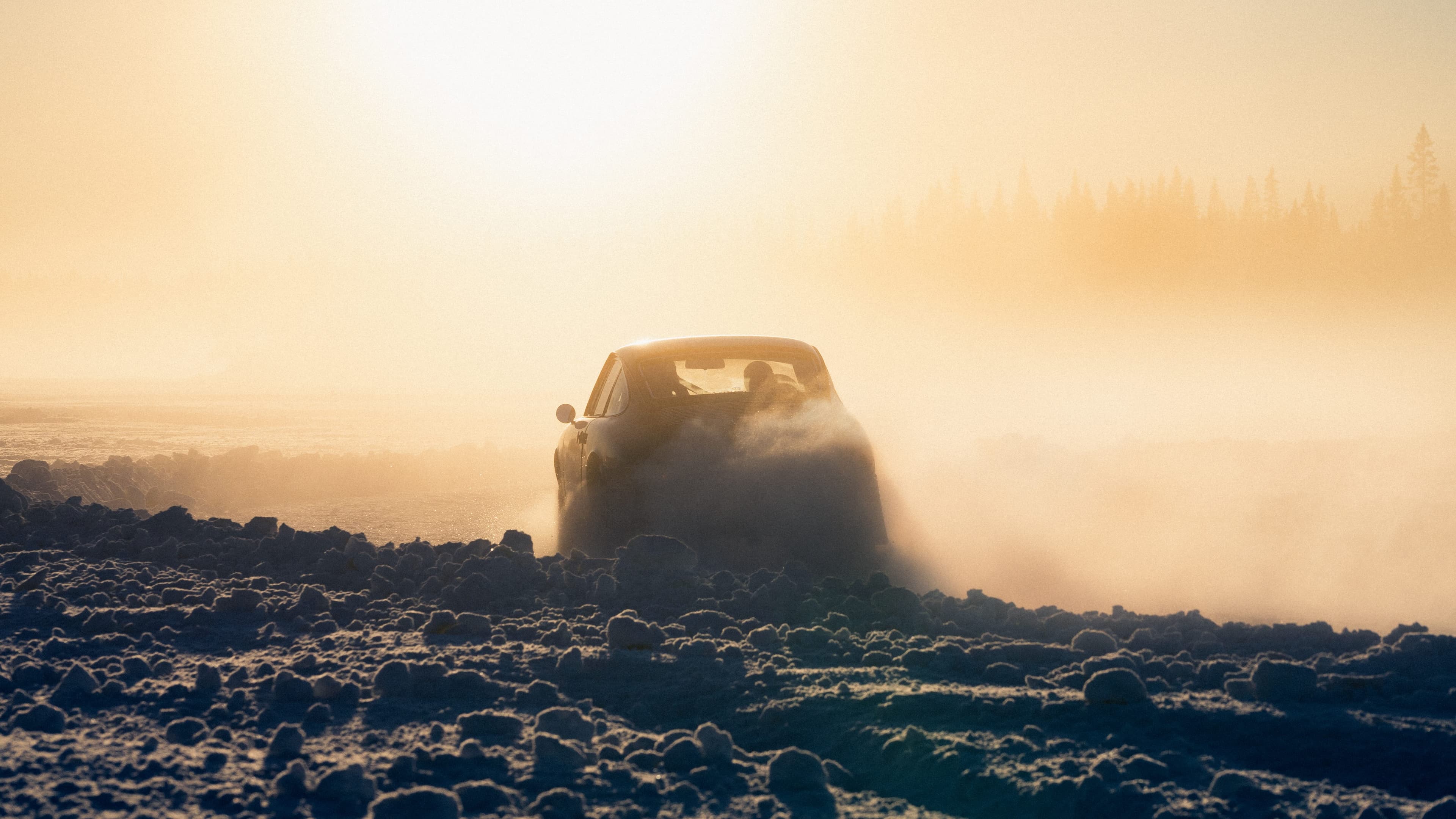 A Porsche 911 drives through a misty, snow-covered landscape at sunset, with hazy sunlight and distant trees silhouetted in the background.